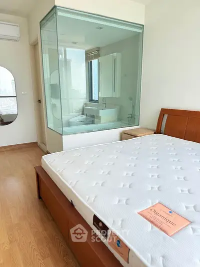 Modern bedroom with glass-enclosed bathroom and city view, featuring wooden flooring and stylish furniture.