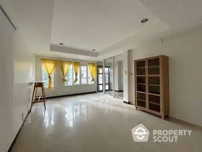 Spacious and well-lit living room with glossy tiled flooring, large windows draped with yellow curtains, and an elegant wooden bookshelf.