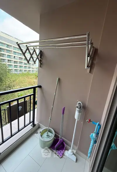 Charming balcony with practical drying rack and cleaning tools, overlooking lush greenery and modern building.