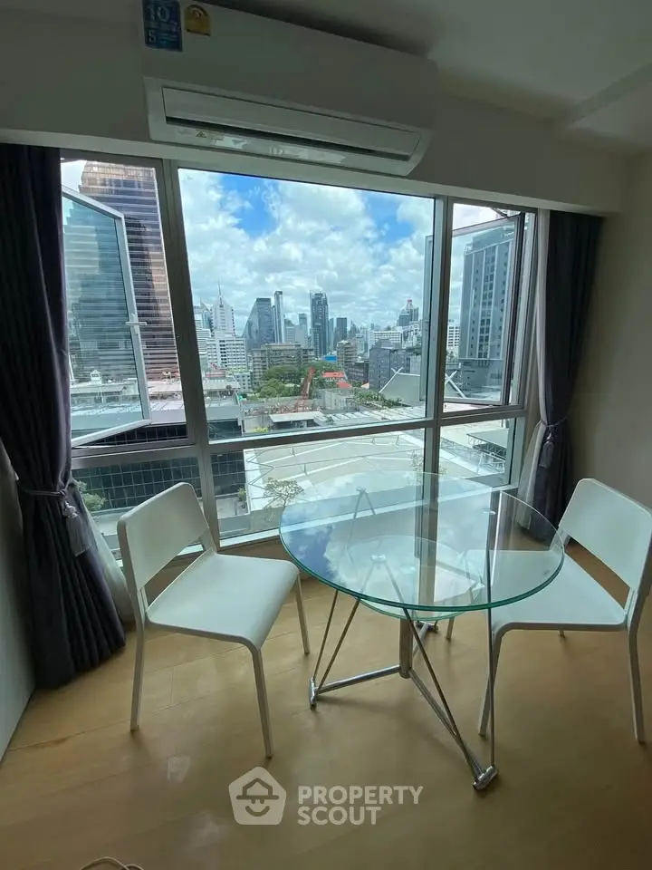 Modern apartment with stunning city view, featuring a glass dining table and chairs by large windows.