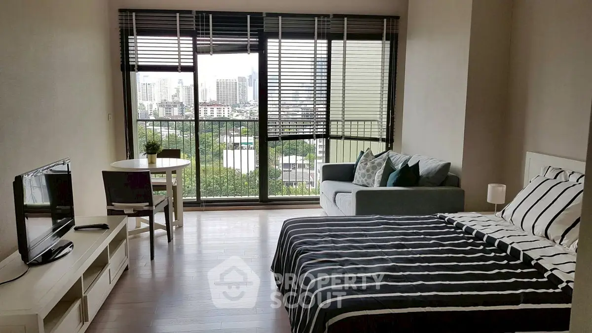 Modern studio apartment with city view, featuring cozy bed, stylish sofa, and natural light through large windows.