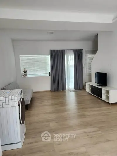 Spacious living room with modern decor and natural light, featuring a washing machine and TV setup.
