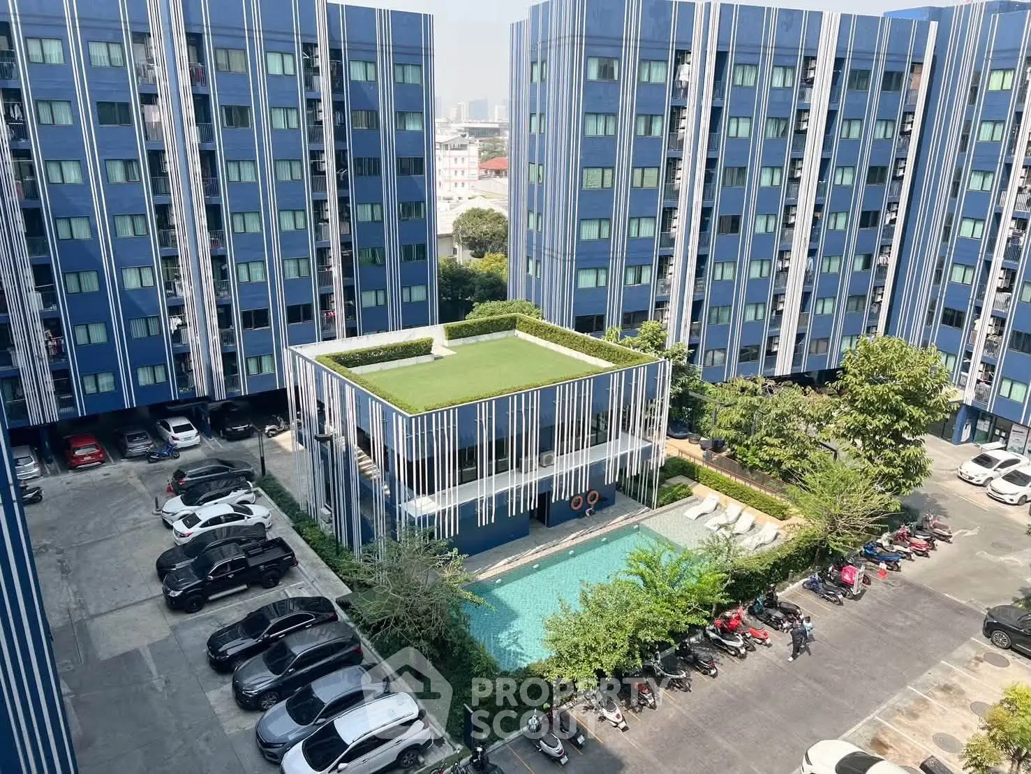 Modern apartment complex with pool and parking area, featuring blue facade and lush greenery.