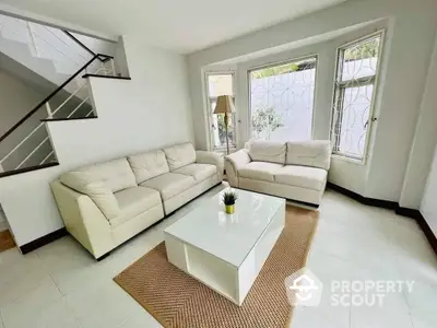 Bright and airy living room with modern white sofas, a chic coffee table, and large windows allowing ample natural light, complemented by a stylish staircase.