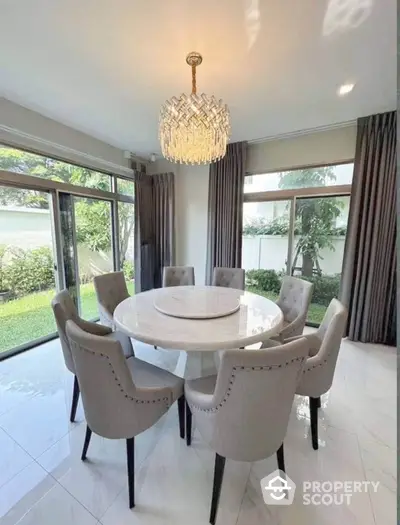 Elegant dining room with round marble table and chandelier, overlooking lush garden views.