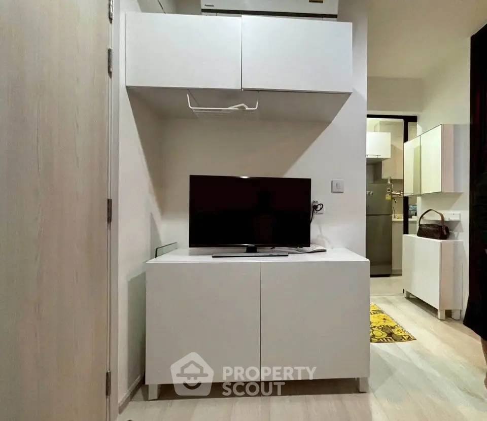 Modern living room with sleek white cabinetry and flat-screen TV, open kitchen in background.