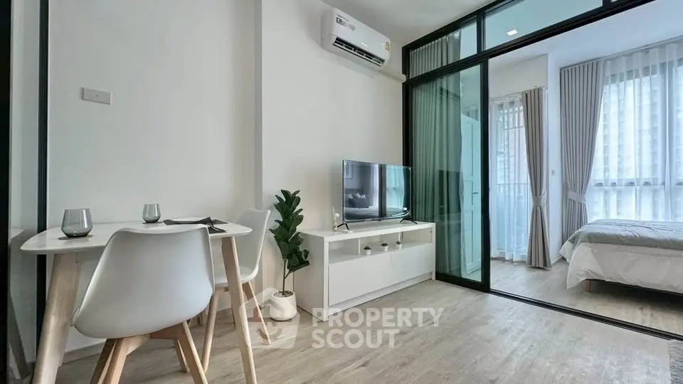 Modern living room with glass partition leading to cozy bedroom, featuring sleek furniture and natural light.