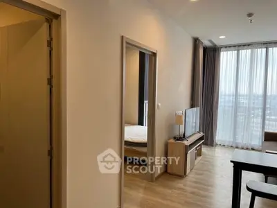 Modern apartment interior with open living room and bedroom view, featuring large windows and wooden flooring.