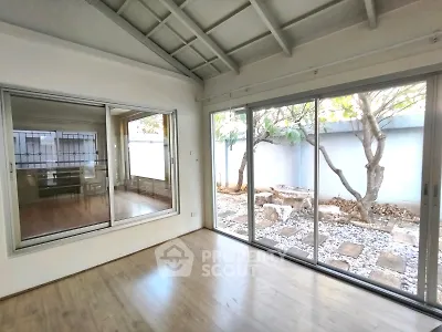 Spacious room with large windows and garden view, featuring wooden flooring and high ceiling.