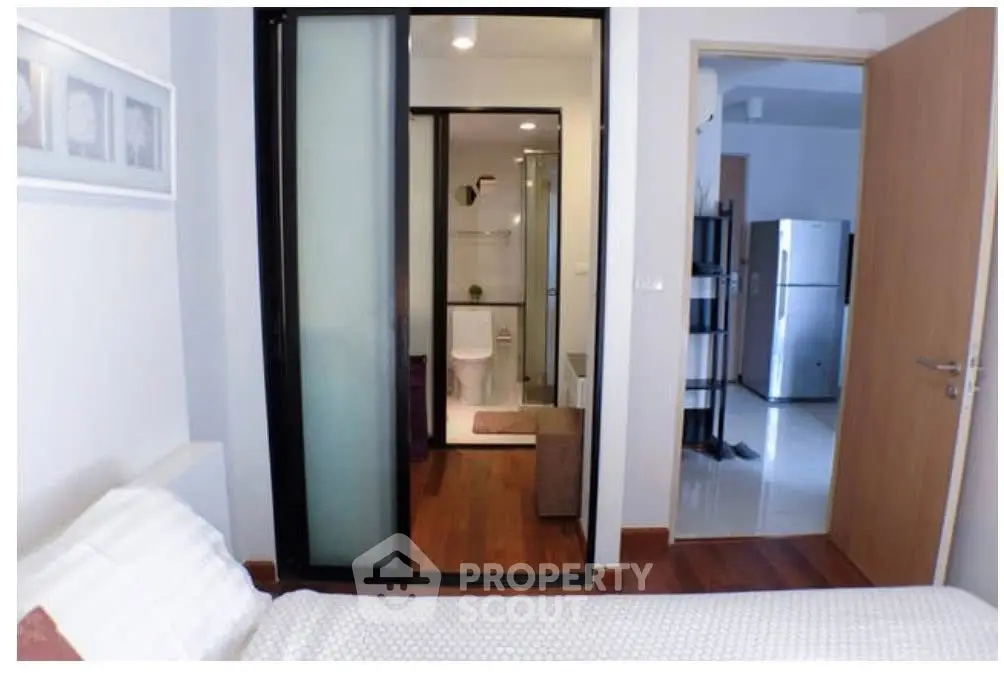 Modern bedroom with ensuite bathroom and view of kitchen area, featuring sleek design and wooden flooring.