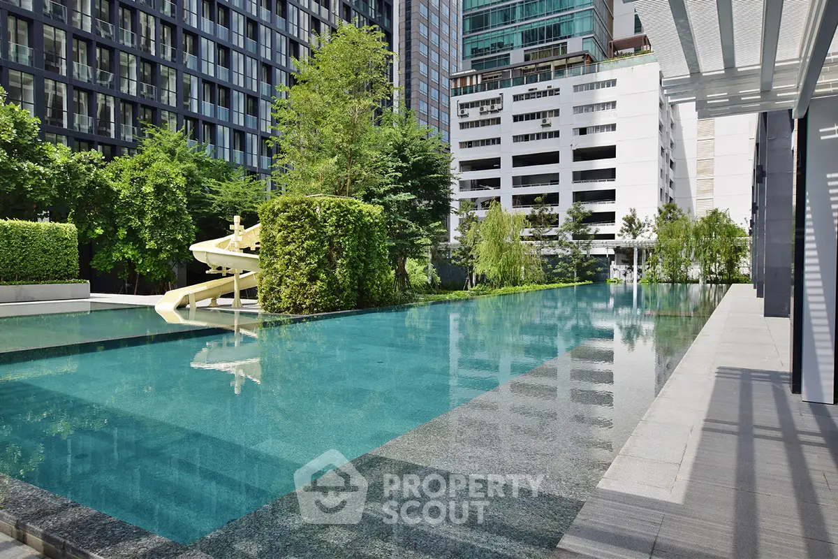 Luxurious urban swimming pool with slide and lush greenery in modern residential complex.