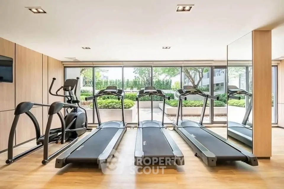Modern gym with treadmills and elliptical machines, large windows offering garden views.