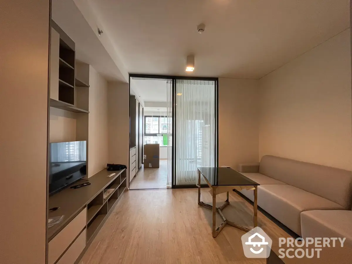 Modern living room with sleek furniture and ample natural light, featuring a large TV and a comfortable L-shaped sofa, leading to a cozy balcony.