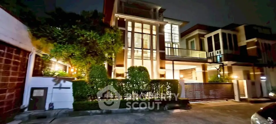 Luxurious modern house exterior with large windows and lush greenery at night.