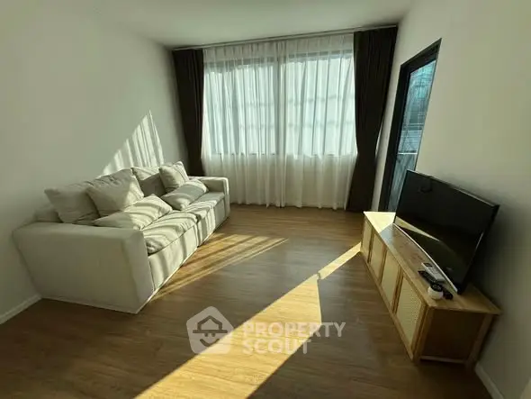 Bright living room with cozy sofa and TV, featuring large windows and wooden flooring.