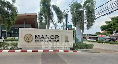 Entrance sign of Manor Sanambinnam with palm trees and street view.