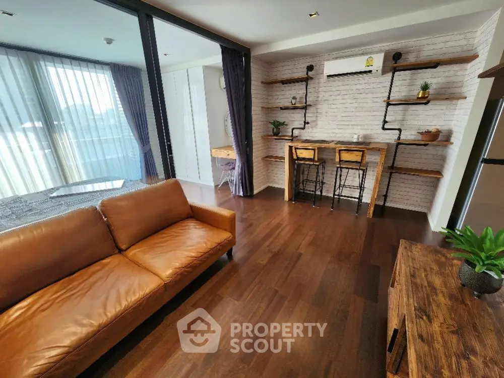 Spacious living room with elegant hardwood flooring, a plush leather sofa, and chic brick accent wall, leading to a sunlit balcony.