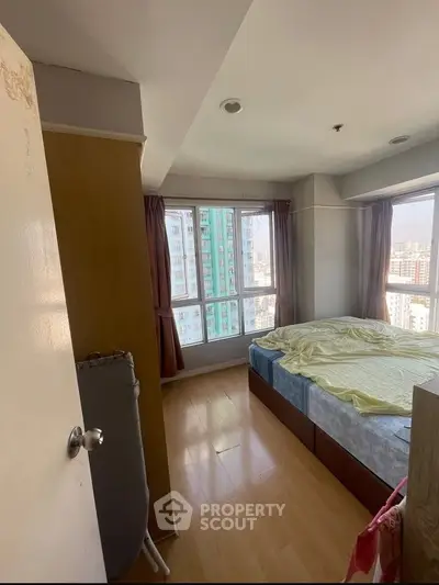 Cozy bedroom with large windows offering city views, featuring wooden flooring and a comfortable bed.