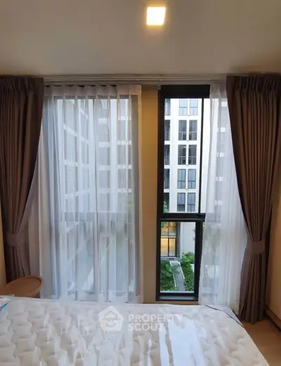 Modern bedroom with large windows offering a city view, featuring elegant curtains and a comfortable mattress.