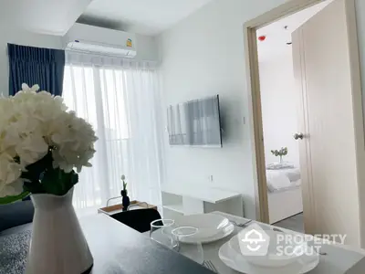 Elegant dining area with modern decor, pristine white tableware, and a bouquet of fresh flowers, complemented by a cozy bedroom glimpse and natural light.