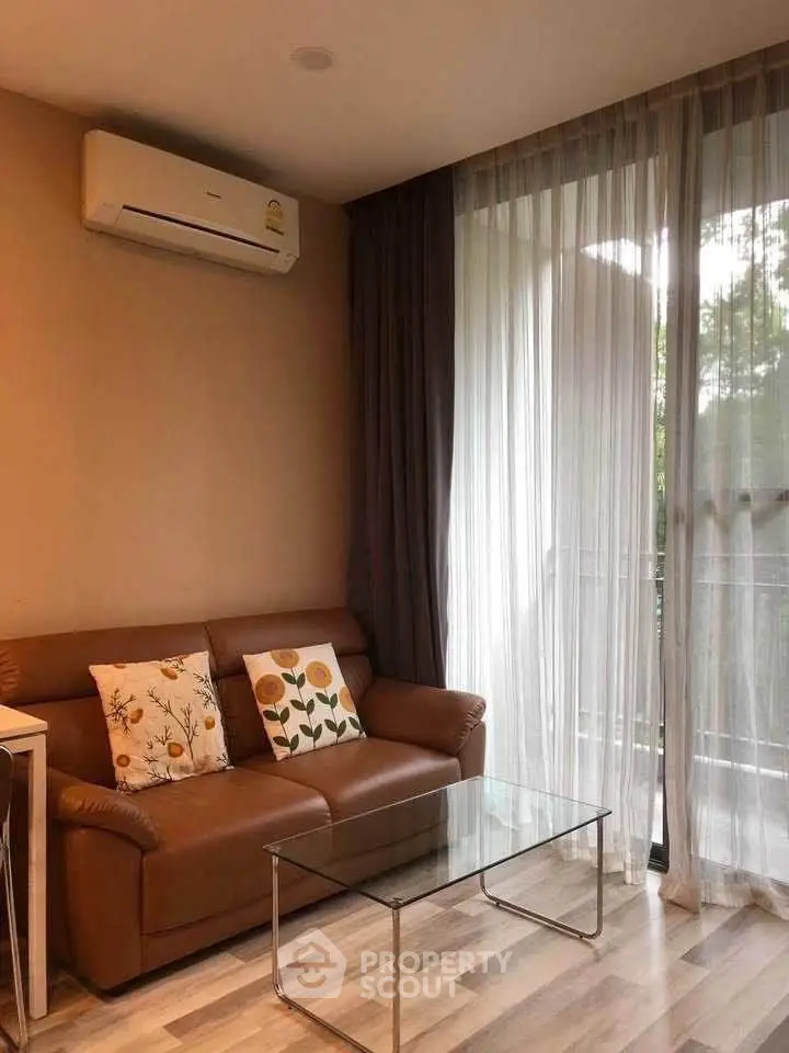 Cozy living room with leather sofa and glass coffee table, featuring large window with sheer curtains.