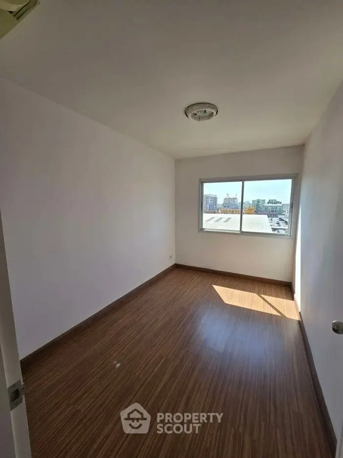 Bright empty room with wooden flooring and large window offering city view.