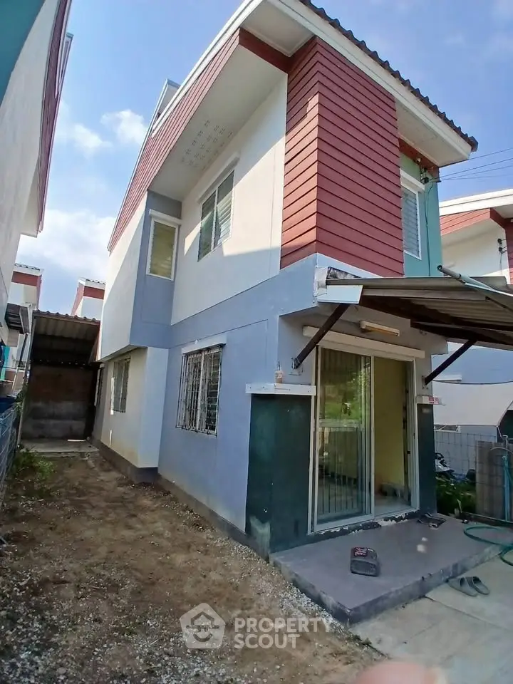 Modern two-story house with red and blue exterior, featuring a small yard and sliding glass doors.