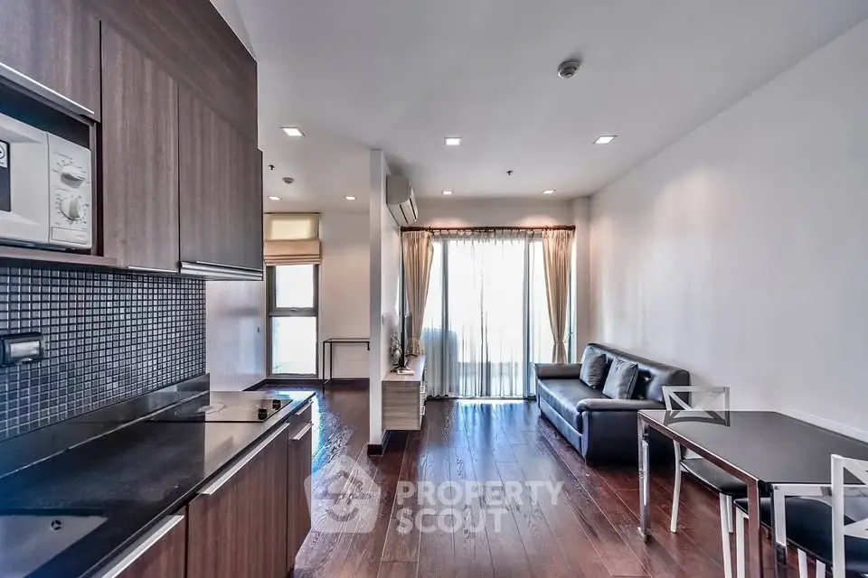Modern living room with sleek kitchen in open layout, featuring hardwood floors and natural light.