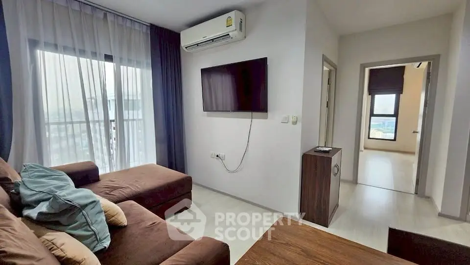 Modern living room with brown sofa, wall-mounted TV, and large window with city view.