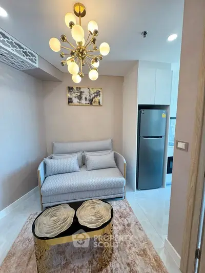 Modern living room with elegant chandelier and cozy sofa, adjacent to a sleek kitchen with stainless steel fridge.