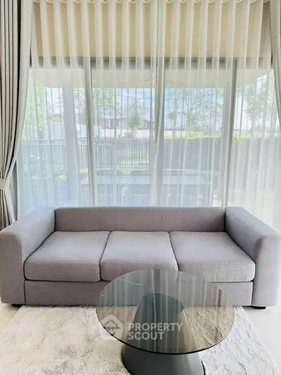 Elegant living room with modern gray sofa and glass coffee table, featuring large windows with sheer curtains.