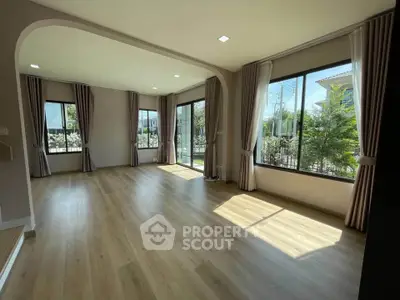 Spacious sunlit living room with large windows and elegant curtains, offering a serene garden view.