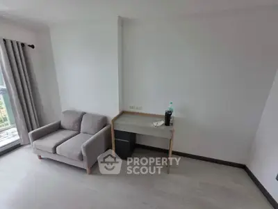 Minimalist living room with gray sofa and small desk, featuring large window with curtains.