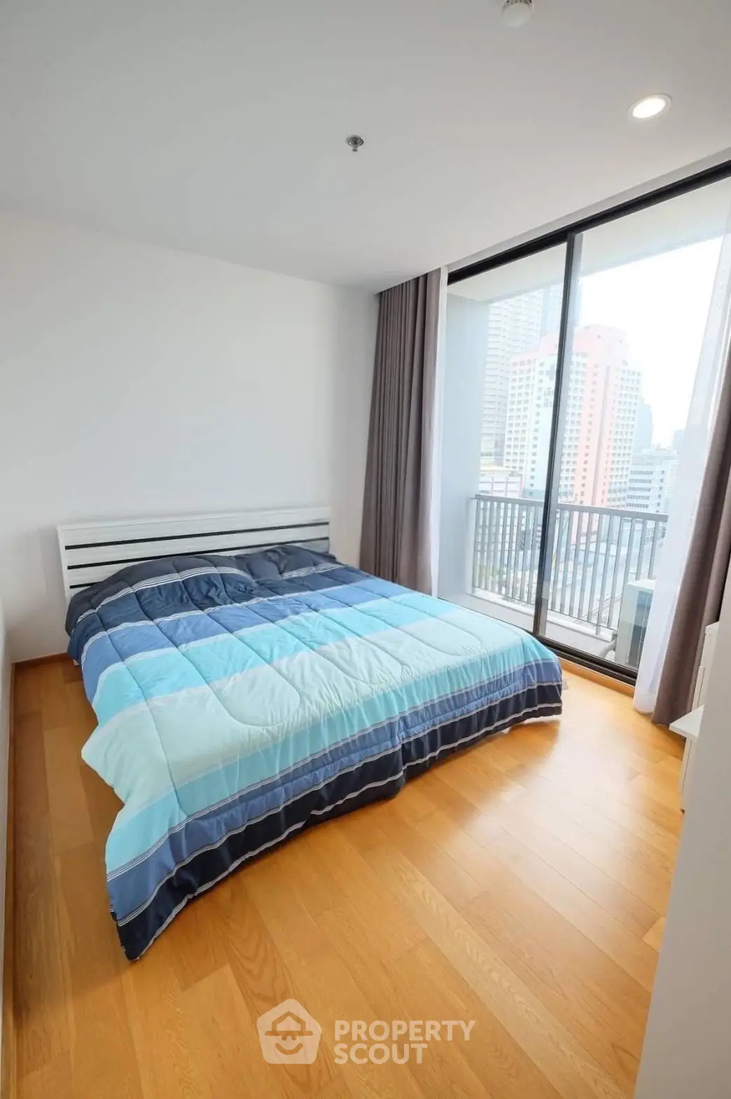 Modern bedroom with large window and city view, featuring a cozy bed and wooden flooring.
