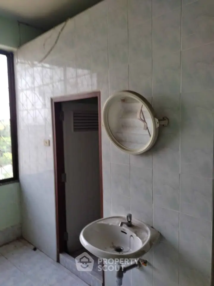 Simple bathroom with wall-mounted sink and round mirror, featuring tiled walls and a small window.