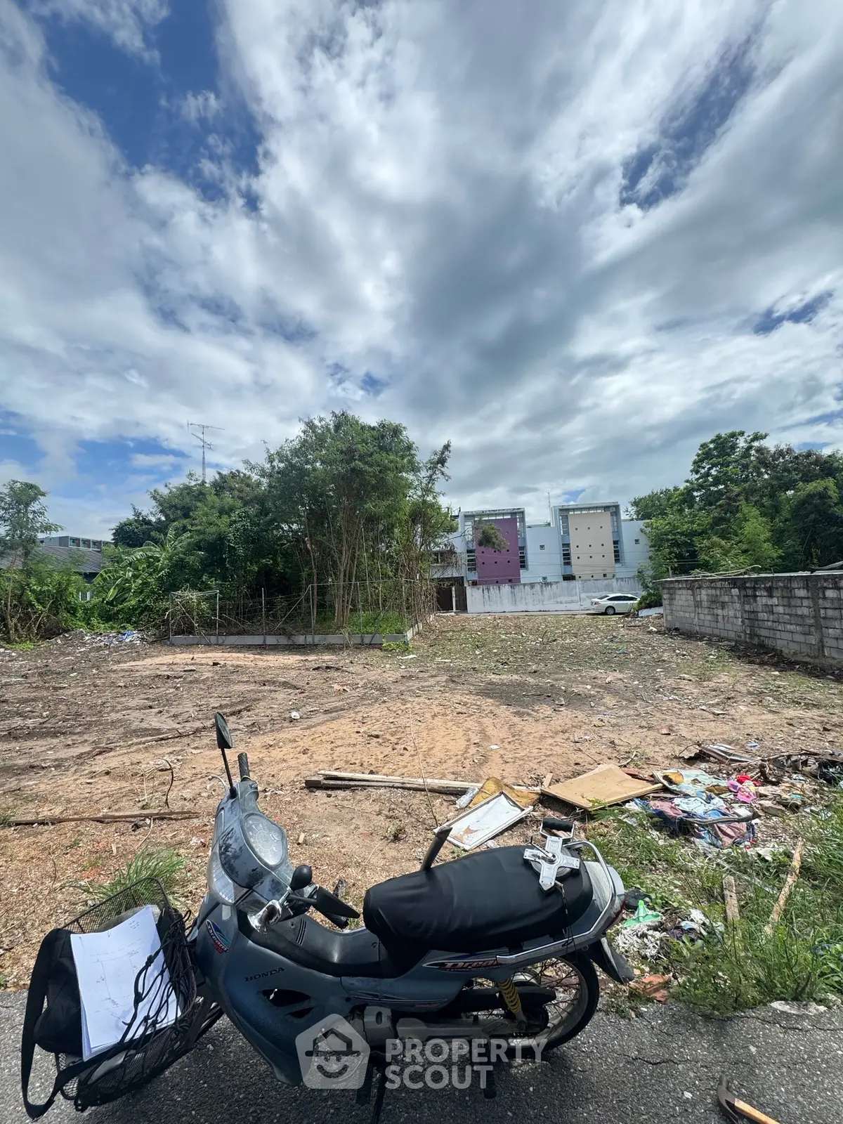 Vacant land plot with potential for development under a cloudy sky, featuring nearby buildings and greenery.