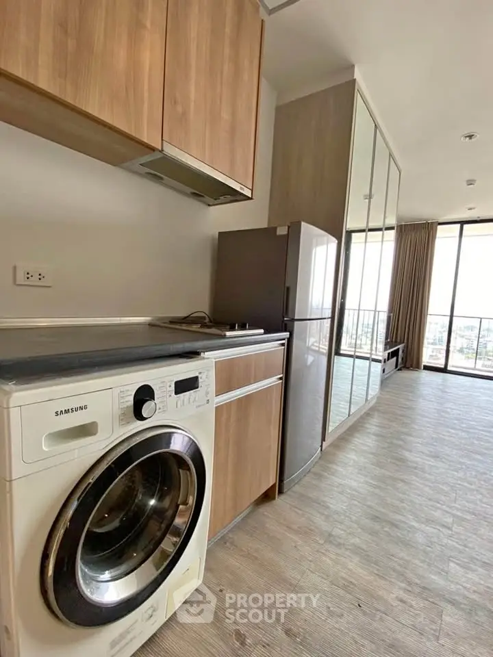 Modern apartment kitchen with washing machine and fridge, featuring sleek wooden cabinets and large windows.