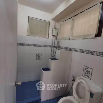 Compact bathroom with tiled shower and toilet, featuring high windows for natural light.