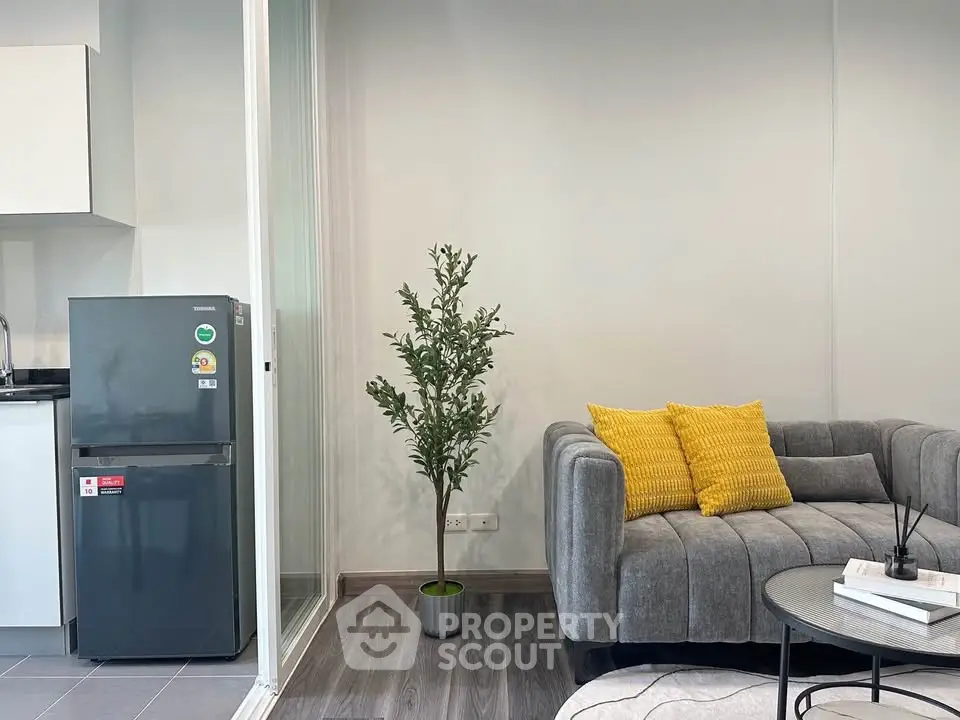 Modern living room with stylish gray sofa, yellow cushions, and compact kitchen area featuring a sleek fridge.