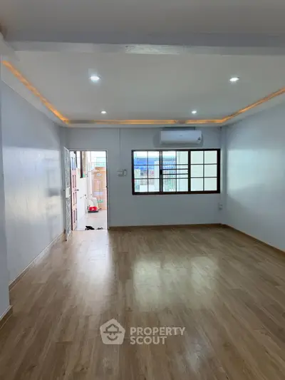 Spacious living room with wooden flooring and large window, featuring modern lighting and air conditioning.