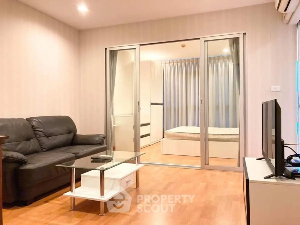 Modern living room with glass partition leading to cozy bedroom, featuring sleek furniture and warm lighting.