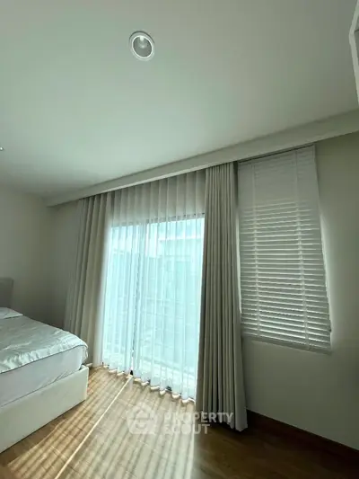 Bright bedroom with large window and sheer curtains, wooden flooring, and cozy bed setup.
