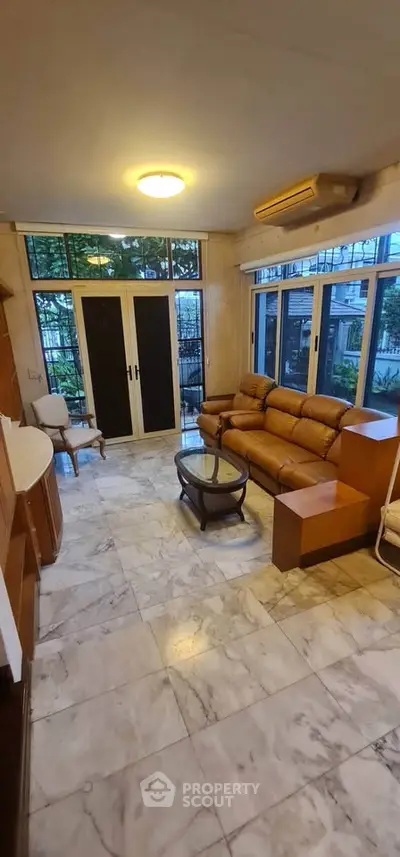 Spacious living room with marble flooring and large windows overlooking a garden, featuring a cozy leather sofa.