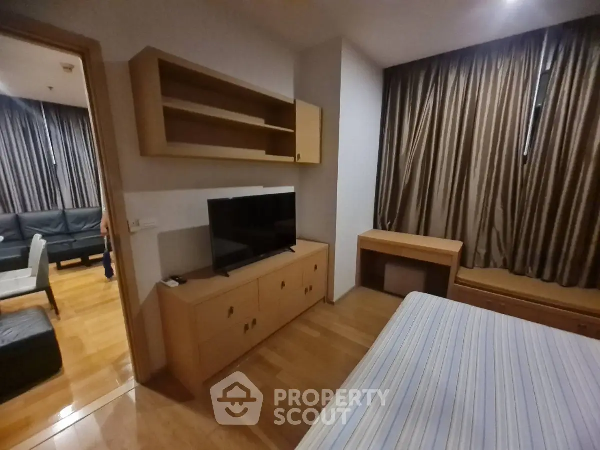 Cozy bedroom with wooden furniture and TV, adjacent to a living room with modern decor.