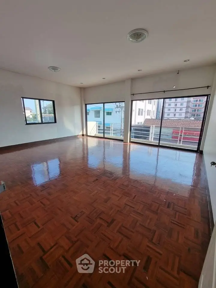 Spacious sunlit room with polished wooden floors and large windows offering city views.