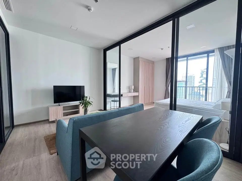 Modern open-plan living room with dining area and bedroom, featuring sleek furniture and large windows.