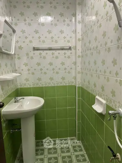 Charming compact bathroom with green and white floral tiles, pedestal sink, and wall-mounted mirror.