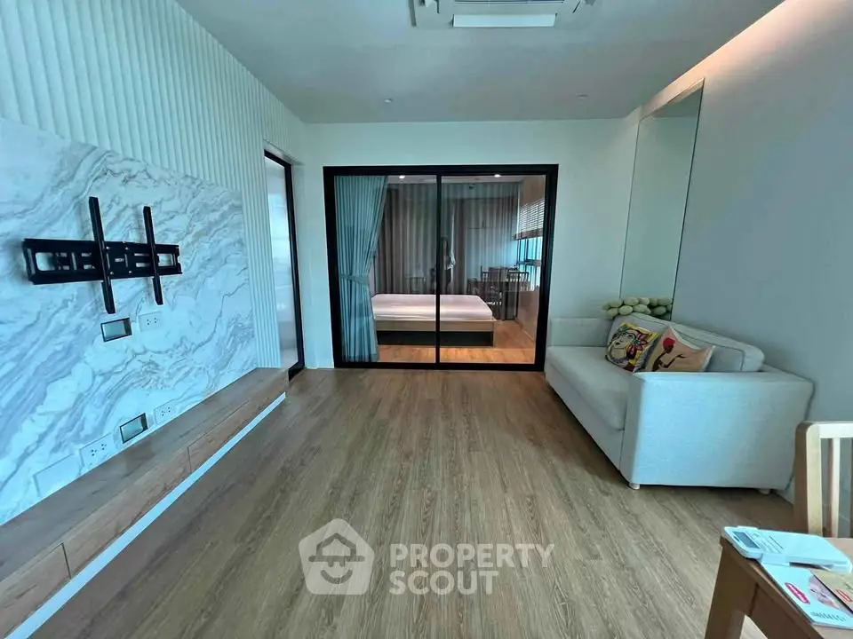 Modern living room with wooden flooring and glass partition leading to bedroom, featuring a cozy sofa and wall mount setup.