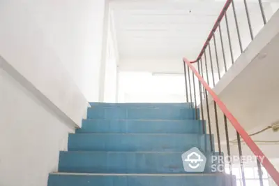 Brightly lit stairway with blue steps and red handrails leading to an airy upper level, offering a glimpse into the building's interior design.