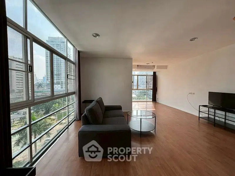 Spacious modern living room with large windows and city view, featuring sleek furniture and wooden flooring.